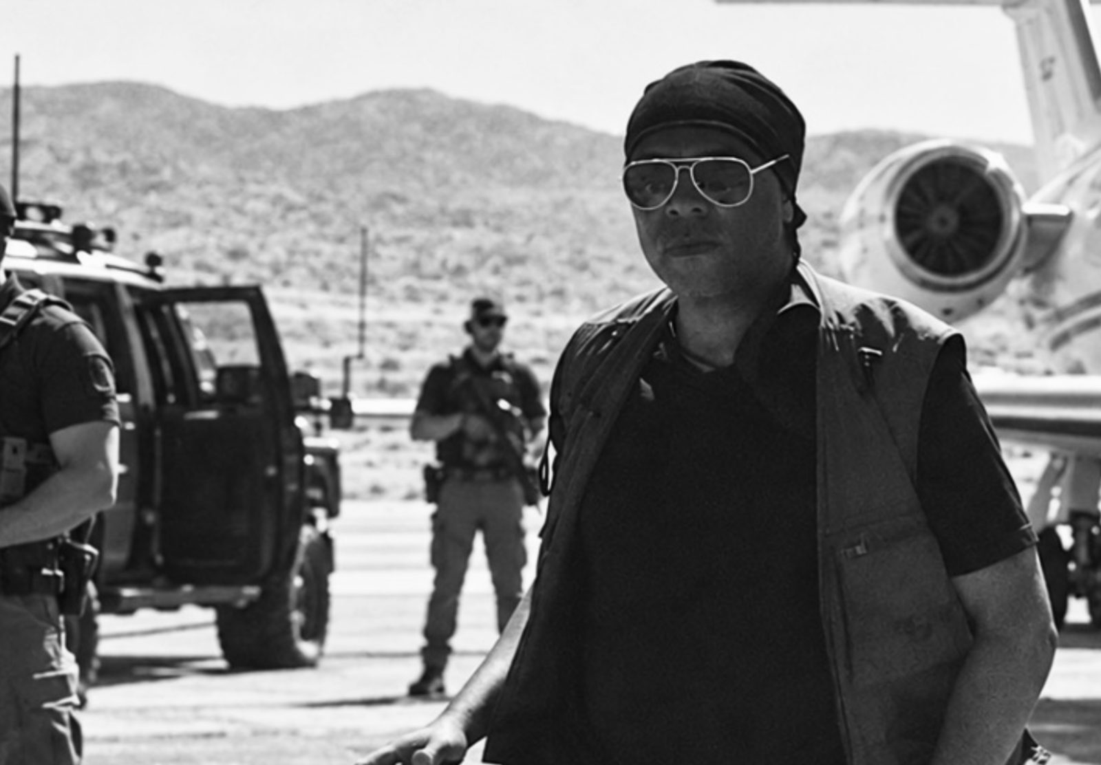 M. Nuri Shakoor in sunglasses and a dark head wrap walking across an airfield, with armed security personnel, a security vehicle, and a private jet behind him